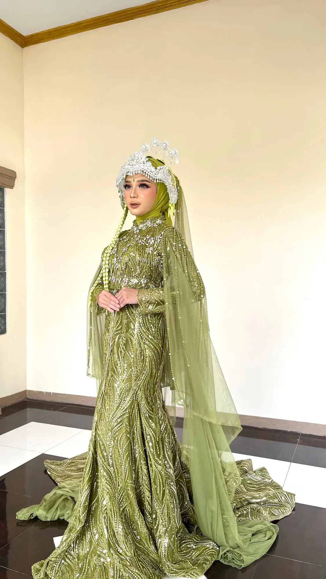 Pengantin bergaun hijau sage dengan jubah tulle dan siger, berdiri di ruang tamu