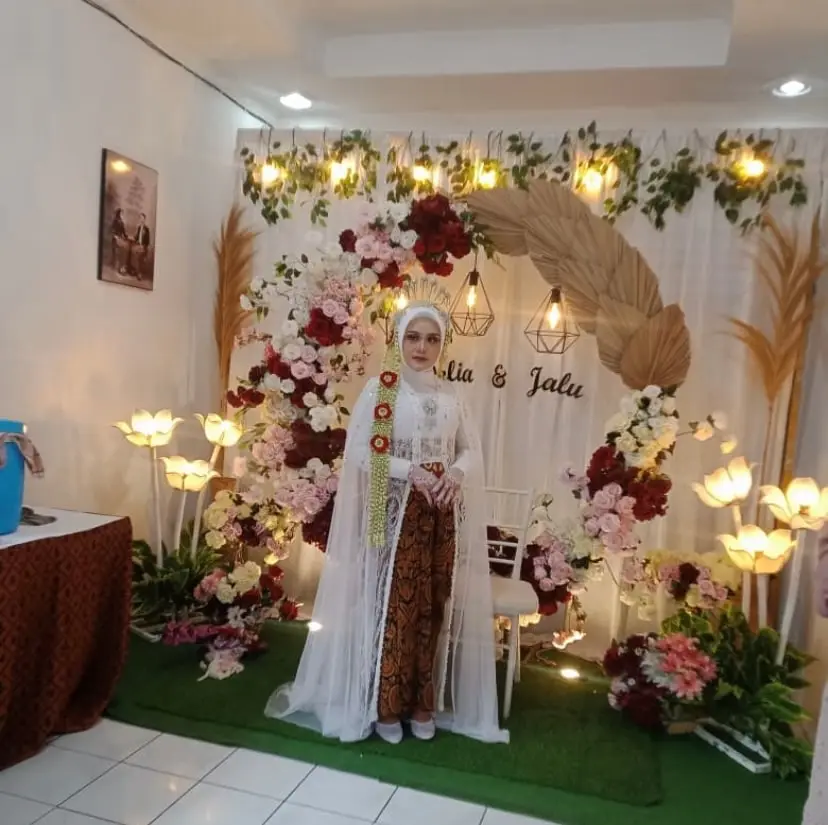Pengantin berkebaya putih dan kain batik di depan dekorasi bunga merah dan putih di rumah