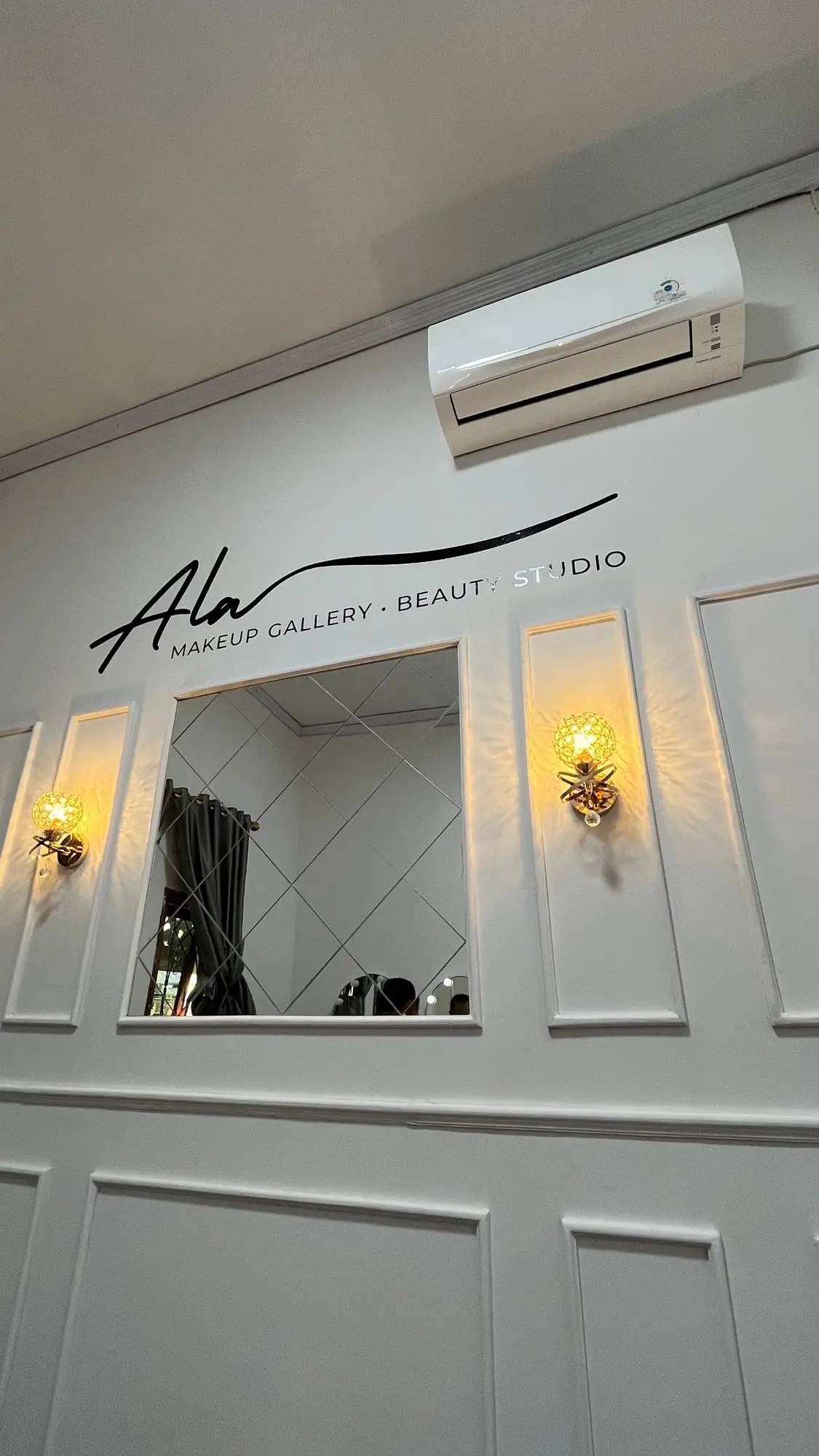 Dinding putih berpanel di studio dengan tulisan Ala Makeup Gallery Beauty Studio, cermin, dan dua lampu dinding